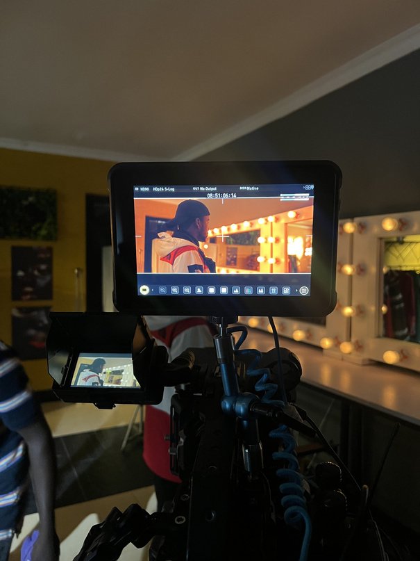 Monitor showing a subject framed against a bulb-lit vanity mirror in a dressing room