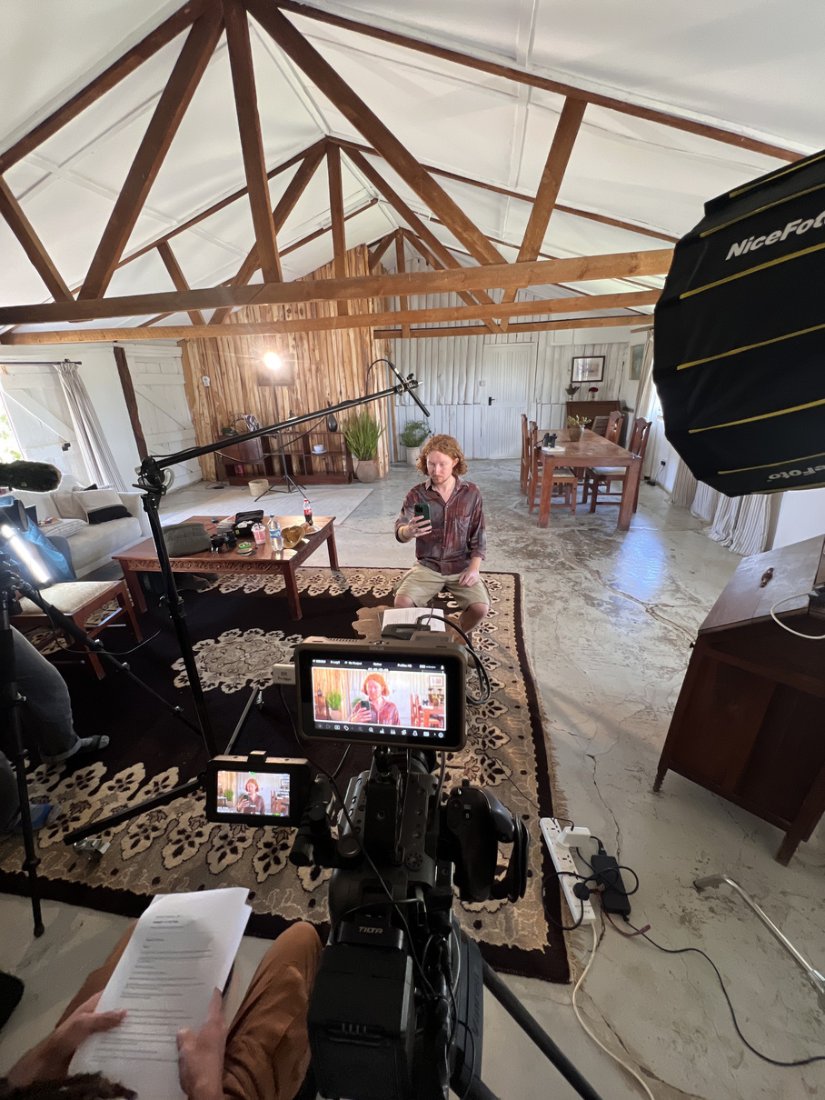 Subject seated on a patterned rug under a timber-trussed roof, softbox at frame right