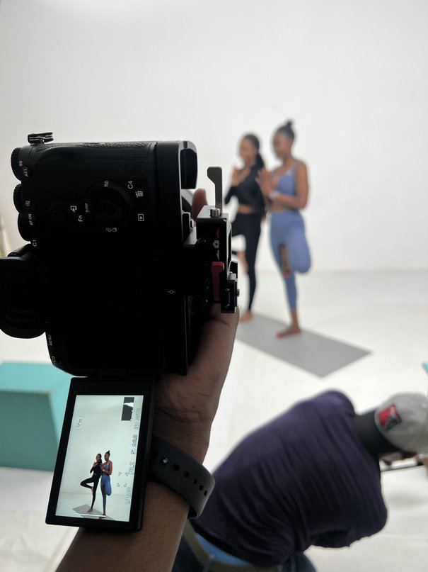 Handheld camera photographing two women in a yoga pose on a white studio backdrop