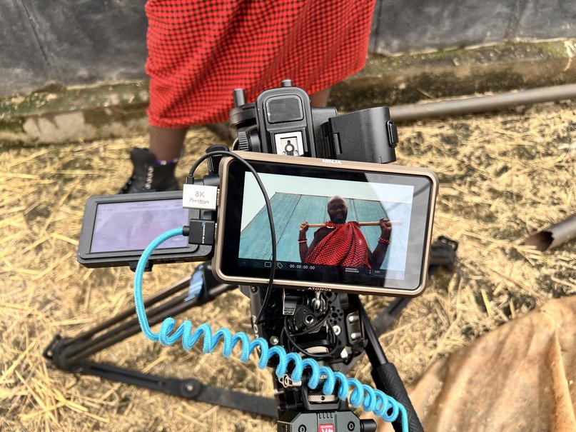Monitor on a tripod showing a man in a red Maasai shuka on straw ground