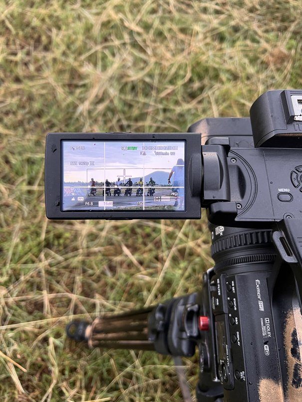 Camcorder flip screen in long grass showing a line of motorcycles below hills